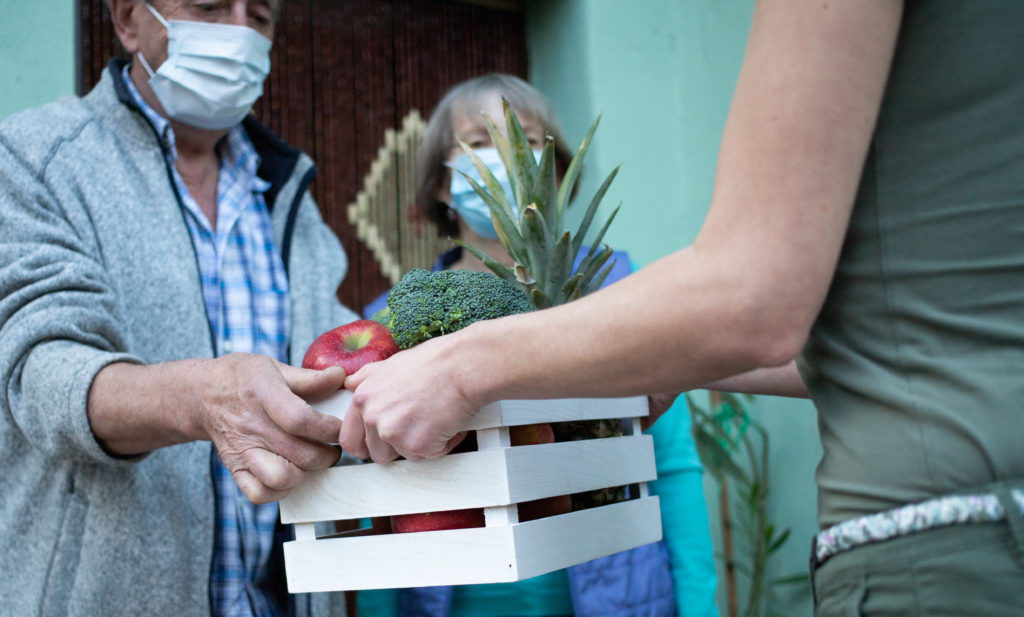 An older couple wearing PPE masks receive the gift of a box of fruit and vegetables from a visitor